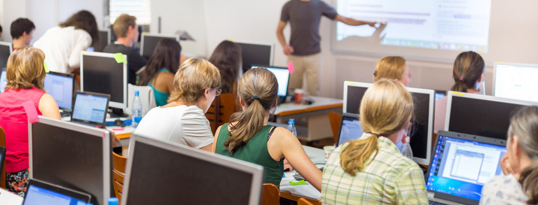 Computerunterricht Viele Menschen sitzen in einem Kursraum vor Computern. Vorne steht ein Lehrer und zeigt etwas auf einem Bildschirm.